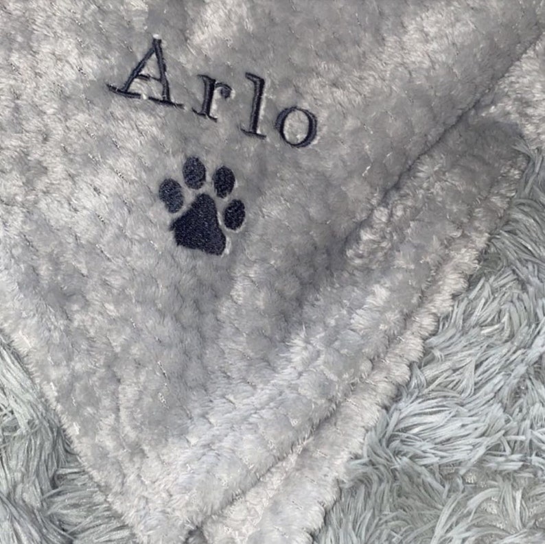 May include: A gray and white plush blanket with the name "Arlo" embroidered on it. A black paw print is embroidered below the name.