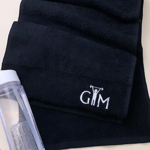 K&ouml;nnte beinhalten: Schwarzes Sporttuch mit wei&szlig; gesticktem Text "GIM" und einer Strichm&auml;nnchenfigur, die Gewichte hebt.