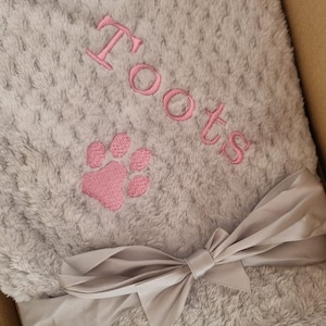 May include: A gray plush blanket with a pink paw print and the name "Toots" embroidered on it. The blanket is tied with a gray satin ribbon.