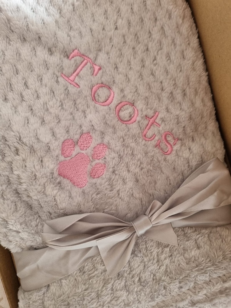 May include: A gray plush blanket with a pink paw print and the name "Toots" embroidered on it. The blanket is tied with a gray satin ribbon.