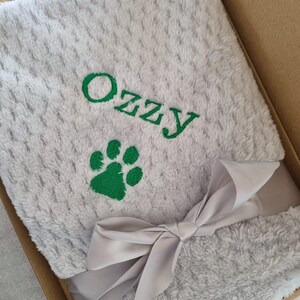 May include: A gray plush blanket with a green paw print and the name "Ozzy" embroidered on it. The blanket is tied with a gray satin ribbon and is in a brown box.