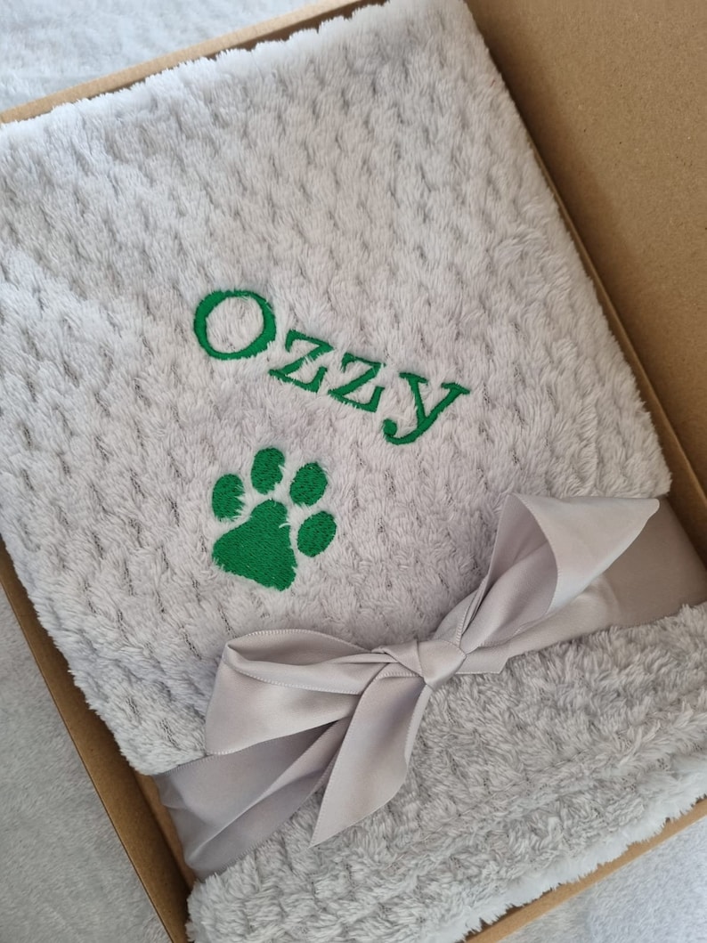 May include: A gray plush blanket with a green paw print and the name "Ozzy" embroidered on it. The blanket is tied with a gray satin ribbon and is in a brown box.