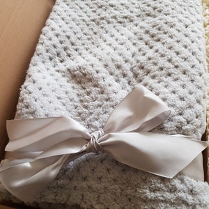 May include: A soft, gray, plush blanket with a white satin ribbon tied in a bow. The blanket is folded and placed in a brown box.