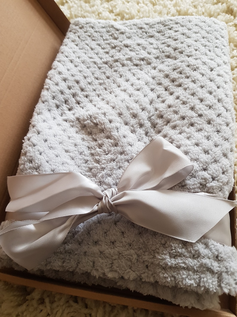 May include: A soft, gray, plush blanket with a white satin ribbon tied in a bow. The blanket is folded and placed in a brown box.