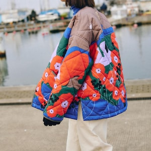 Peut inclure: Une femme portant un bonnet noir et une veste matelassée colorée à motif floral. La veste présente des fleurs rouges, bleues, vertes et blanches. La femme porte un pantalon blanc.