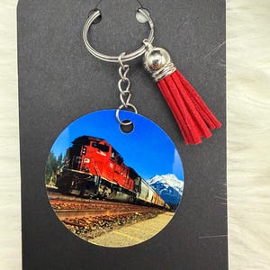 May include: Round keychain featuring a vibrant image of a red train against a backdrop of mountains and a clear blue sky. The keychain has a silver ring, chain, and a red tassel. A travel-themed accessory.