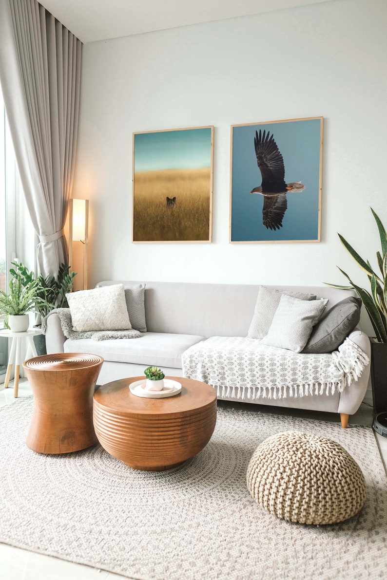 Op de afbeelding: Een woonkamer met een grijze bank, twee ingelijste natuurprints en houten salontafels. De prints tonen een vos in een veld en een vliegende adelaar. Een geweven vloerkleed en een gebreide poef vullen de ruimte aan.
