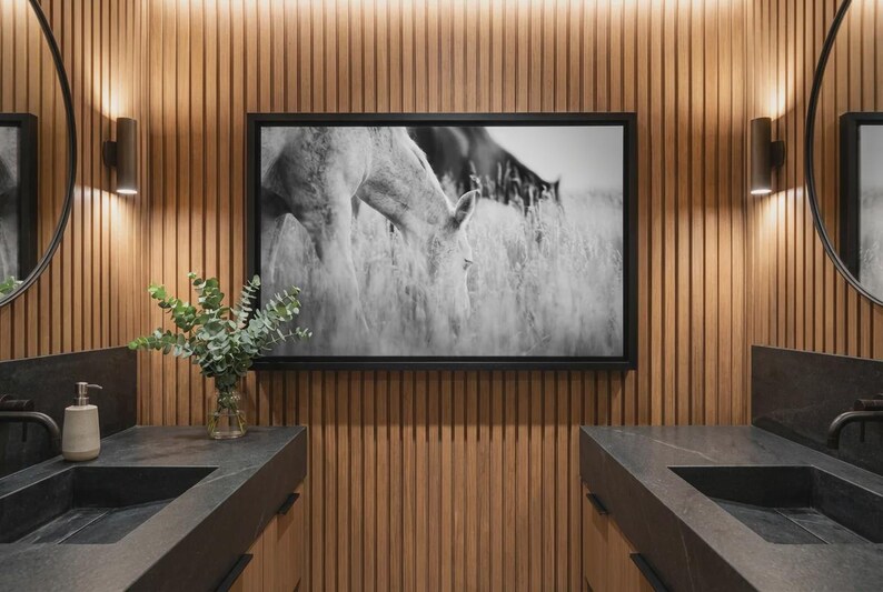 Pu&ograve; includere: Un bagno moderno con una fotografia incorniciata in bianco e nero di cavalli al pascolo in un campo. Le pareti sono rivestite in legno e ci sono due lavandini neri con specchi e applique abbinati. Un vaso di verde &egrave; posto sul piano di lavoro.