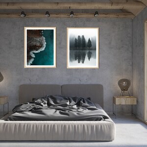 May include: Two framed photographs hanging on a gray wall above a gray bed with a gray duvet. The photo on the left shows an aerial view of a turquoise ocean with white waves crashing on a rocky shore. The photo on the right shows a black and white image of a group of trees reflected in a still body of water.