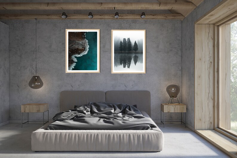 May include: Two framed photographs hanging on a gray wall above a gray bed with a gray duvet. The photo on the left shows an aerial view of a turquoise ocean with white waves crashing on a rocky shore. The photo on the right shows a black and white image of a group of trees reflected in a still body of water.