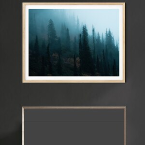 May include: A framed landscape print featuring a misty forest scene. The artwork showcases tall, dark green trees shrouded in fog, with a light blue sky peeking through. The frame is light-colored wood. Below the artwork is a dark gray cabinet.
