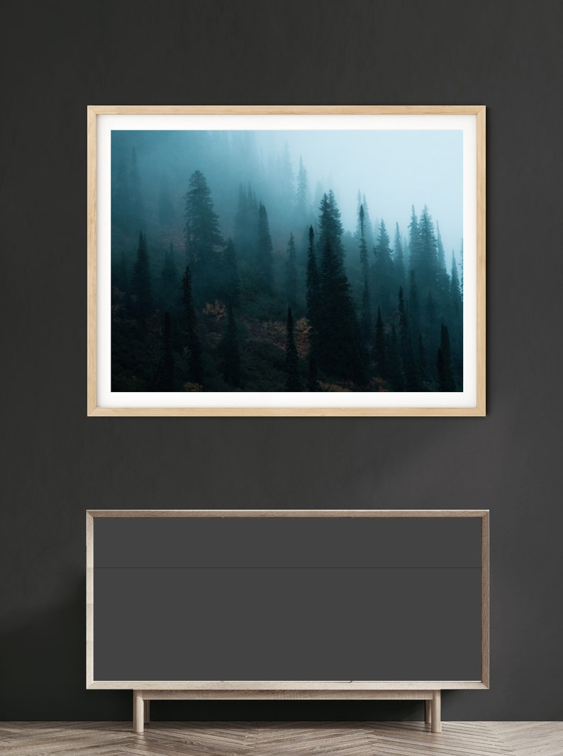 May include: A framed landscape print featuring a misty forest scene. The artwork showcases tall, dark green trees shrouded in fog, with a light blue sky peeking through. The frame is light-colored wood. Below the artwork is a dark gray cabinet.