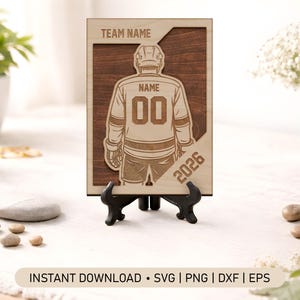 Peut inclure: Plaque en bois sur le thème du hockey, représentant un maillot de joueur avec le numéro "00" et l'année "2026". La plaque est présentée sur un support noir. Les mots "TEAM NAME" et "NAME" sont également visibles.