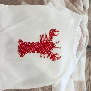 Pode incluir: Body branco com uma aplicação de lagosta vermelha. O desenho da lagosta apresenta um padrão de pontos e é costurado na frente. O body é feito de tecido branco macio.