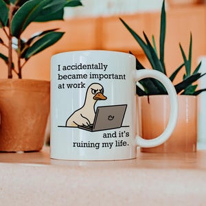 Op de afbeelding: Witte keramische mok met een humoristisch ontwerp met een cartoon eend die op een laptop werkt. De tekst luidt: "Ik ben per ongeluk belangrijk geworden op het werk en het verpest mijn leven."