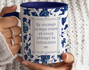 May include: White ceramic mug with a blue interior and handle. The mug features a blue floral pattern and a framed quote that reads, "My courage always rises at every attempt to intimidate me."