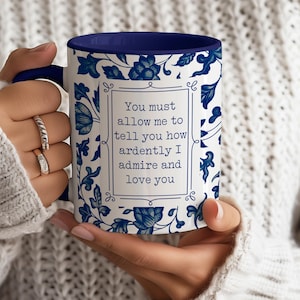 Può includere: Tazza in ceramica bianca con un motivo floreale blu e interno blu. La tazza presenta la citazione: "Devi permettermi di dirti quanto ti ammiro e ti amo". La tazza è tenuta da una persona.