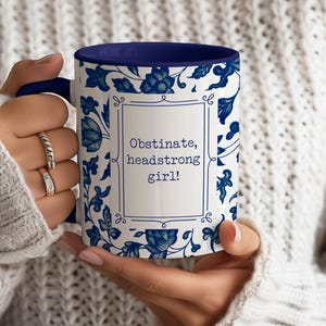 May include: White ceramic mug with a blue floral pattern and a blue rim. The mug has the text "Obstinate, headstrong girl!" in a black frame.