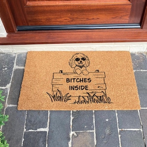 May include: A rectangular doormat made of natural brown coir fiber. The mat features a cartoon dog wearing sunglasses above a sign that reads "BITCHES INSIDE" in black lettering. The design includes a fence and grass.