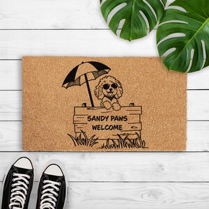 Op de afbeelding: Een rechthoekige bruine kokosmat met een zwart-witte illustratie van een hond met een zonnebril onder een parasol. De tekst "SANDY PAWS WELCOME" staat eronder. Zwarte sneakers staan onderaan.