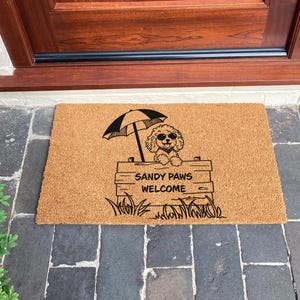 Op de afbeelding: Een rechthoekige deurmat van natuurlijke vezels. Het heeft een zwarte afbeelding van een hond onder een parasol, met de tekst "SANDY PAWS WELCOME" in zwart. De mat ligt voor een houten deur.