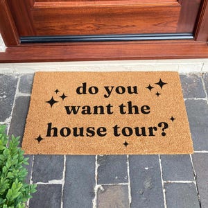 Può includere: Un zerbino rettangolare in fibra di cocco naturale. Il tappeto presenta la frase "do you want the house tour?" in lettere nere, con accenti a forma di stella. Il tappeto è posto davanti a una porta di legno.