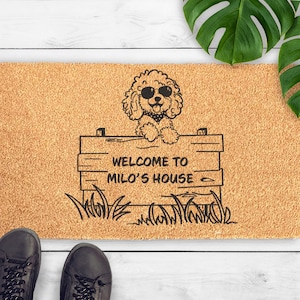 May include: A rectangular coir doormat with a dog illustration wearing sunglasses and a bandana, above a sign that reads "WELCOME TO MILO'S HOUSE". The mat is tan with black text and graphics.