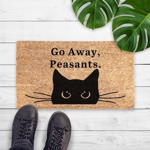 Puede incluir: Felpudo rectangular de fibra de coco natural. Presenta un diseño de cara de gato negro y el texto "Go Away, Peasants." El felpudo está sobre una superficie de madera blanca, con dos pies y hojas verdes visibles.