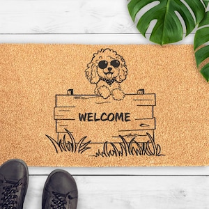 May include: A rectangular doormat with a light brown, textured surface. A black and white illustration of a poodle wearing sunglasses and a bandana is above a wooden sign that reads "WELCOME". The mat is on a white wooden surface, with two black shoes at the bottom.