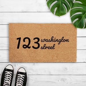 May include: A rectangular tan coir doormat featuring the black printed text 123 washington street. The mat has a coarse, fibrous texture and is positioned horizontally. The numbers 123 are in a large font, followed by the text washington street.