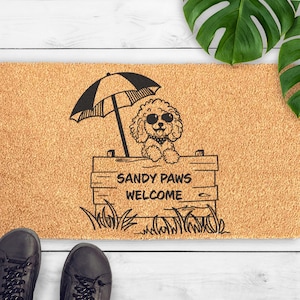 Op de afbeelding: Een rechthoekige deurmat met een lichtbruine achtergrond heeft een zwarte illustratie van een poedel met een zonnebril onder een parasol. De tekst "SANDY PAWS WELCOME" staat onder de hond. Zwarte schoenen staan onderaan.