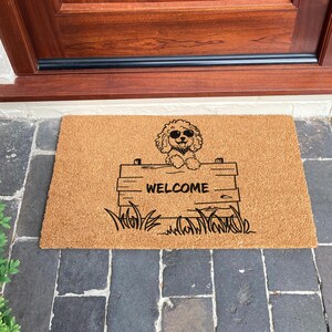 May include: A rectangular doormat made of natural fibers. It features a black graphic of a dog wearing sunglasses, peeking over a wooden sign that reads "WELCOME". The dog is surrounded by grass.