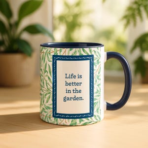 May include: A white ceramic mug with a dark blue handle and interior rim. The exterior features a green leaf pattern surrounding a central rectangular frame with a dark blue border. The text inside reads: Life is better in the garden.