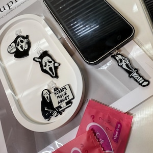 May include: A black smartphone with a Scream-themed phone charm, a white tray with Scream-themed pins, and two pink mystery bags. The pins feature the Scream mask and text. The bags say "mystery bag" and "Open it up."