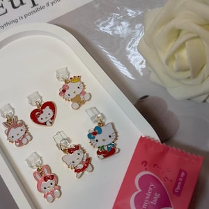 Puede incluir: Una colección de pendientes de esmalte de colores con personajes de dibujos animados, incluyendo Hello Kitty y un conejito, expuestos en una bandeja blanca. También hay una bolsa misteriosa rosa y una rosa blanca. La palabra "Euphoria" es visible en el fondo.