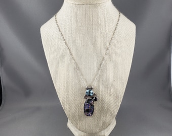 May include: A silver chain necklace with a pendant featuring a purple and blue stone with a smaller blue stone and a black bead.