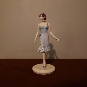 May include: A porcelain figurine of a woman in a blue and white dress with a gold trim. She is standing on a white base.