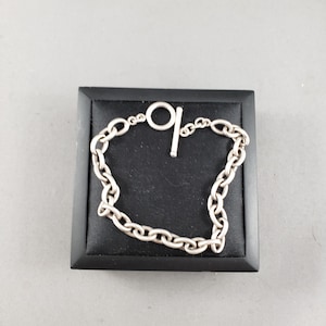 May include: A silver chain bracelet with a toggle clasp, presented on a black square platform. The bracelet comprises interconnected oval links. The toggle clasp has a circular ring and a straight bar. The bracelet is a piece of jewellery.