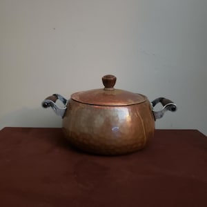 Op de afbeelding: Een koperen pot met een deksel en twee handvatten. De pot heeft een gehamerde textuur en een houten knop op het deksel. De handvatten zijn zilverkleurig met houten grepen. De pot staat op een donkerrode ondergrond.