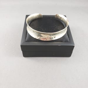 May include: A silver-toned cuff bracelet with a wide, concave band and a hammered, reflective surface texture. The bracelet features an open design, allowing it to be slipped onto the wrist, and is displayed resting on a black square jewellery box.