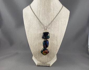 May include: A silver chain necklace with a pendant made of three stacked, square-shaped pieces of iridescent glass. The top piece is black, the middle piece is blue and black, and the bottom piece is multi-colored with red, orange, yellow, and green.