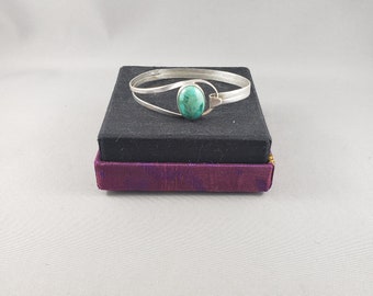 May include: A silver bracelet with a turquoise stone set in the centre. The bracelet is on a black and purple velvet box.