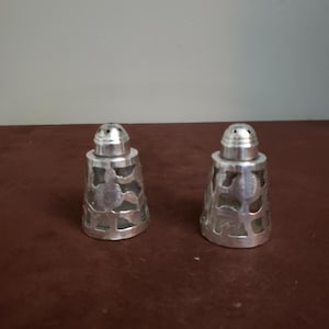 Könnte beinhalten: Zwei silberne Salz- und Pfefferstreuer. Jeder Streuer hat ein dekoratives durchbrochenes Design und eine abgerundete Oberseite mit kleinen Löchern. Die Streuer sind aus glänzendem Metall gefertigt und stehen auf einer braunen Oberfläche.