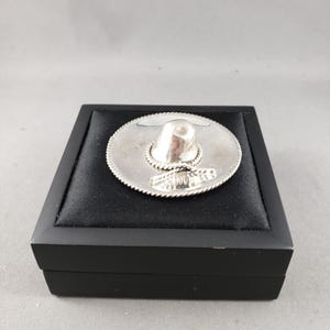 May include: A miniature silver-coloured sombrero pin featuring a smooth crown and a wide, flat brim with a twisted rope-style edge detail. The small hat accessory is displayed resting on a square black velvet-lined presentation box.