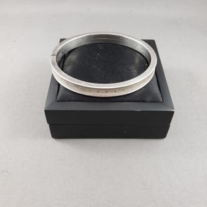 May include: A silver-toned metal bangle bracelet featuring a continuous geometric Greek key pattern engraved along the outer band. The bracelet has a hinged opening mechanism and a smooth, polished finish, resting on a black display stand.