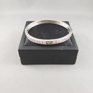 May include: A silver-toned hinged bangle bracelet featuring a row of square-cut light pink stones. The bracelet includes a single rectangular accent piece set with small dark grey stones, providing a contrast to the pink stones along the band.