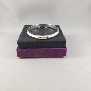May include: A silver bangle bracelet with a gold band and a textured design. The bracelet is displayed on a purple and black box.