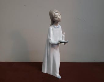 May include: A porcelain figurine of a young person in a white gown, holding a candle on a small silver tray. The figurine has a serene expression and is in a standing pose. The gown has a light blue trim.