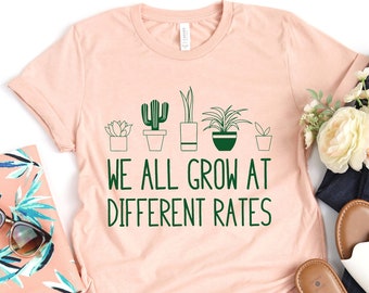 May include: A peach-colored t-shirt with a green graphic of potted plants and the text "We all grow at different rates".