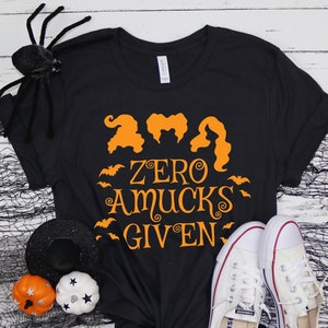 May include: Black t-shirt with orange text that reads "And Zero A-Mucks Given" with a graphic of three witches with orange hair and bats.